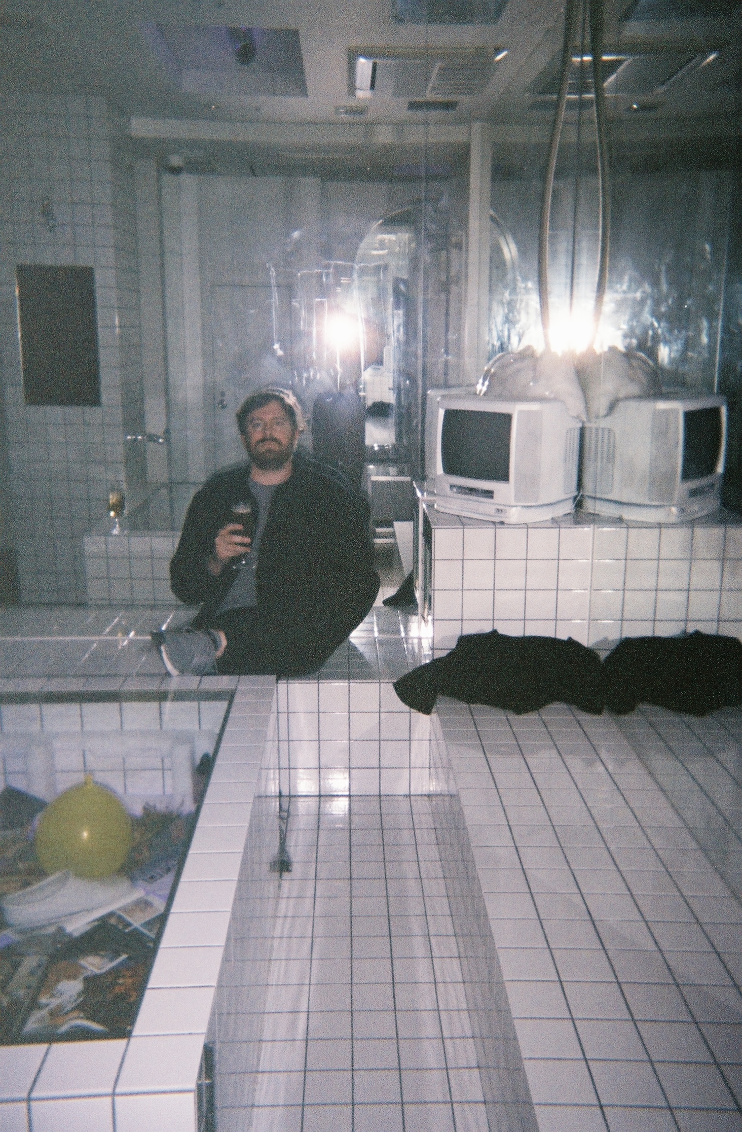 Color film print of a man, holding a drink, sitting in a white tiled area next to two old computers in front of a mirror reflecting the camera flash and obscuring the photographer