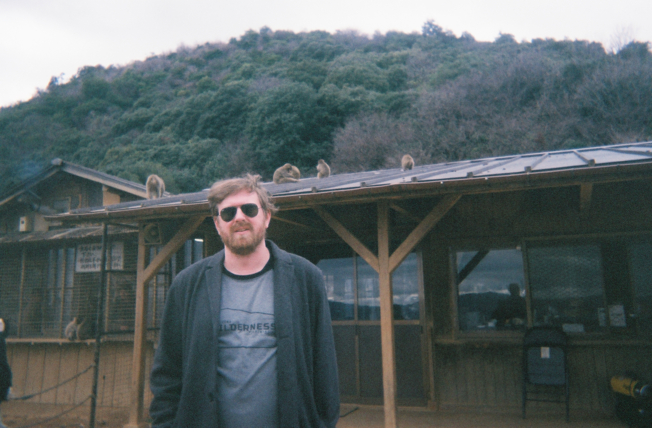 Color film photo of a man in sunglasses standing in front of a structure with macaque monkeys