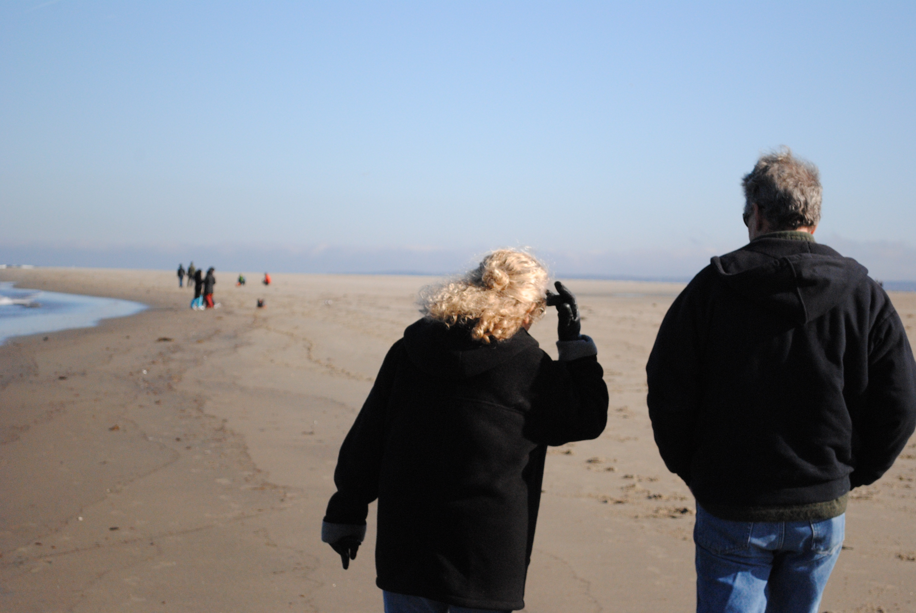 Color photo of a beach and blue sky with a bundled up couple in the foreground walking away from the camera
