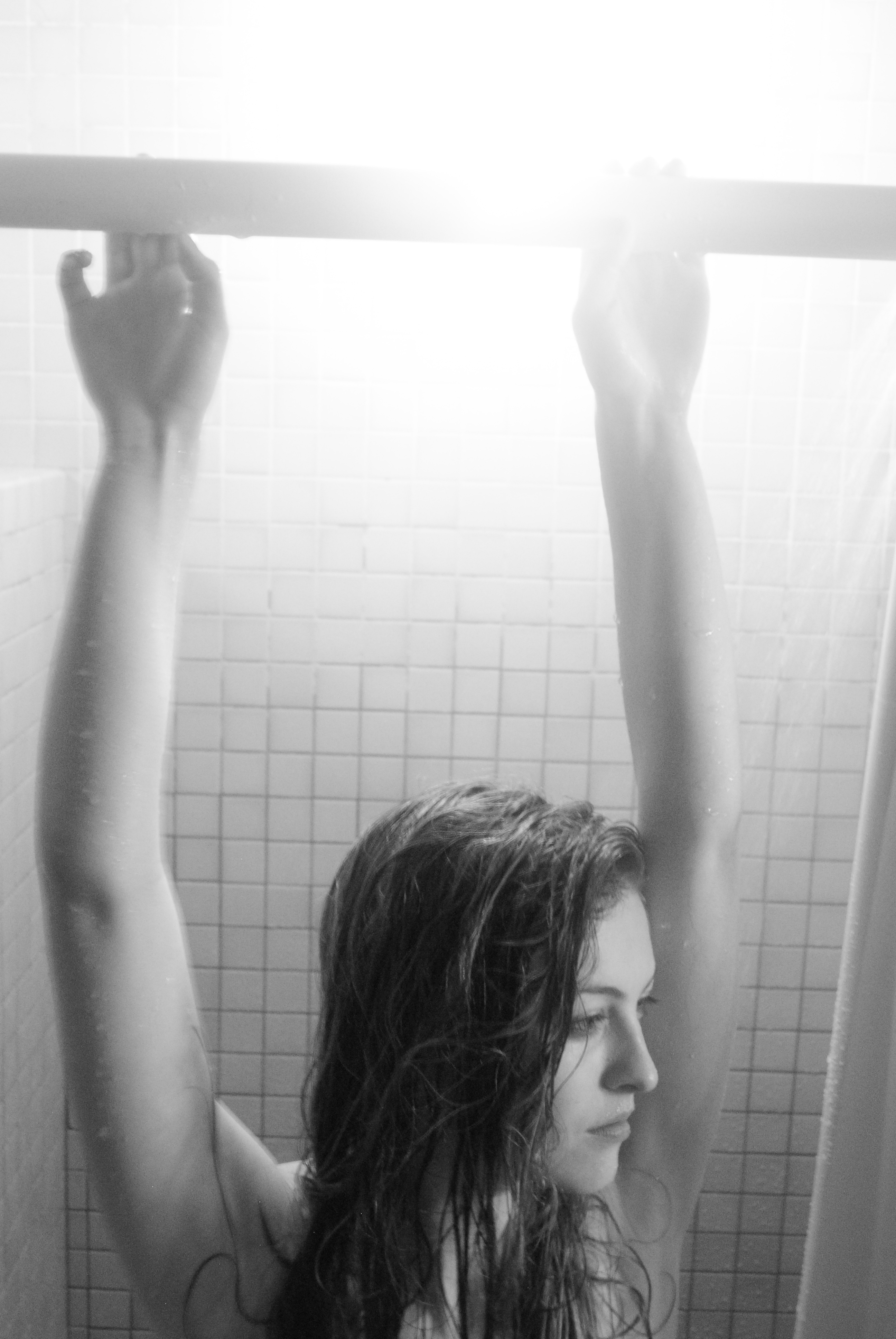Black and white image of a girl from the collarbones up with wet hair standing in front of a shower with her arms raised to hold the curtain bar