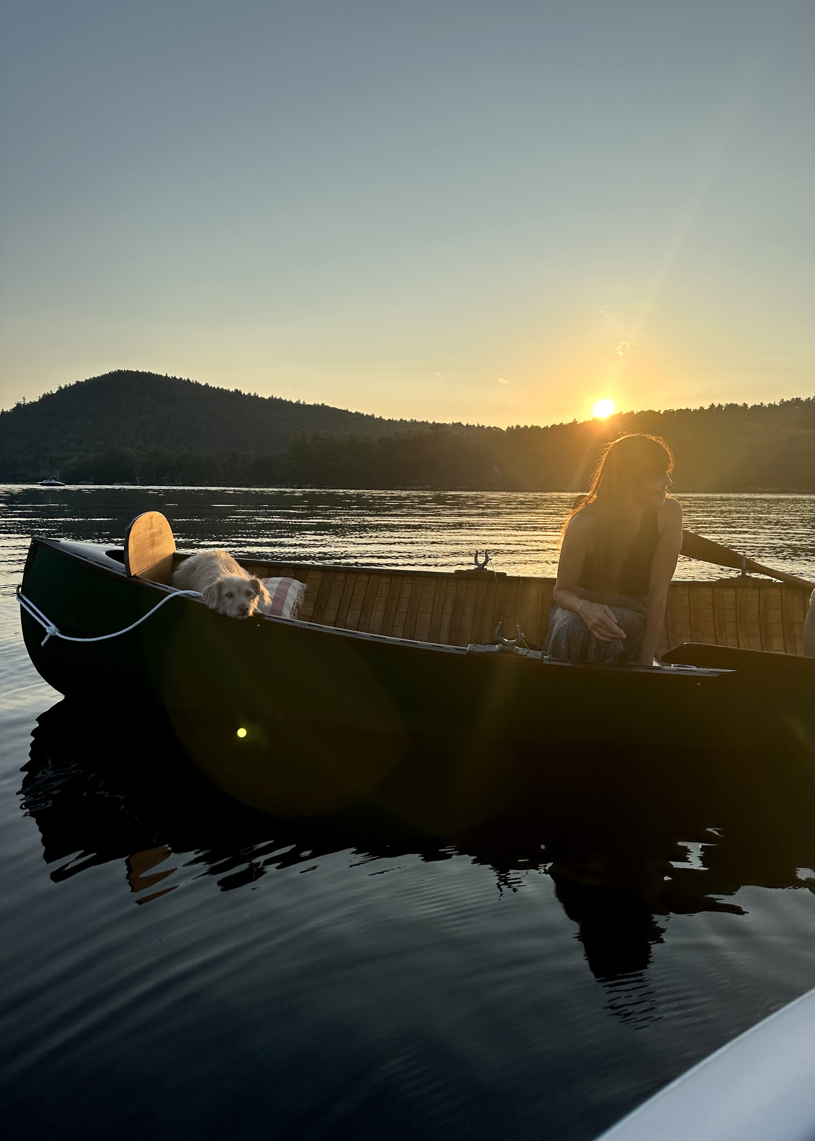 Color photo of a sunset behind mountains in the background and a canoe with a woman and a dog in the foreground with a light flare from the sunset