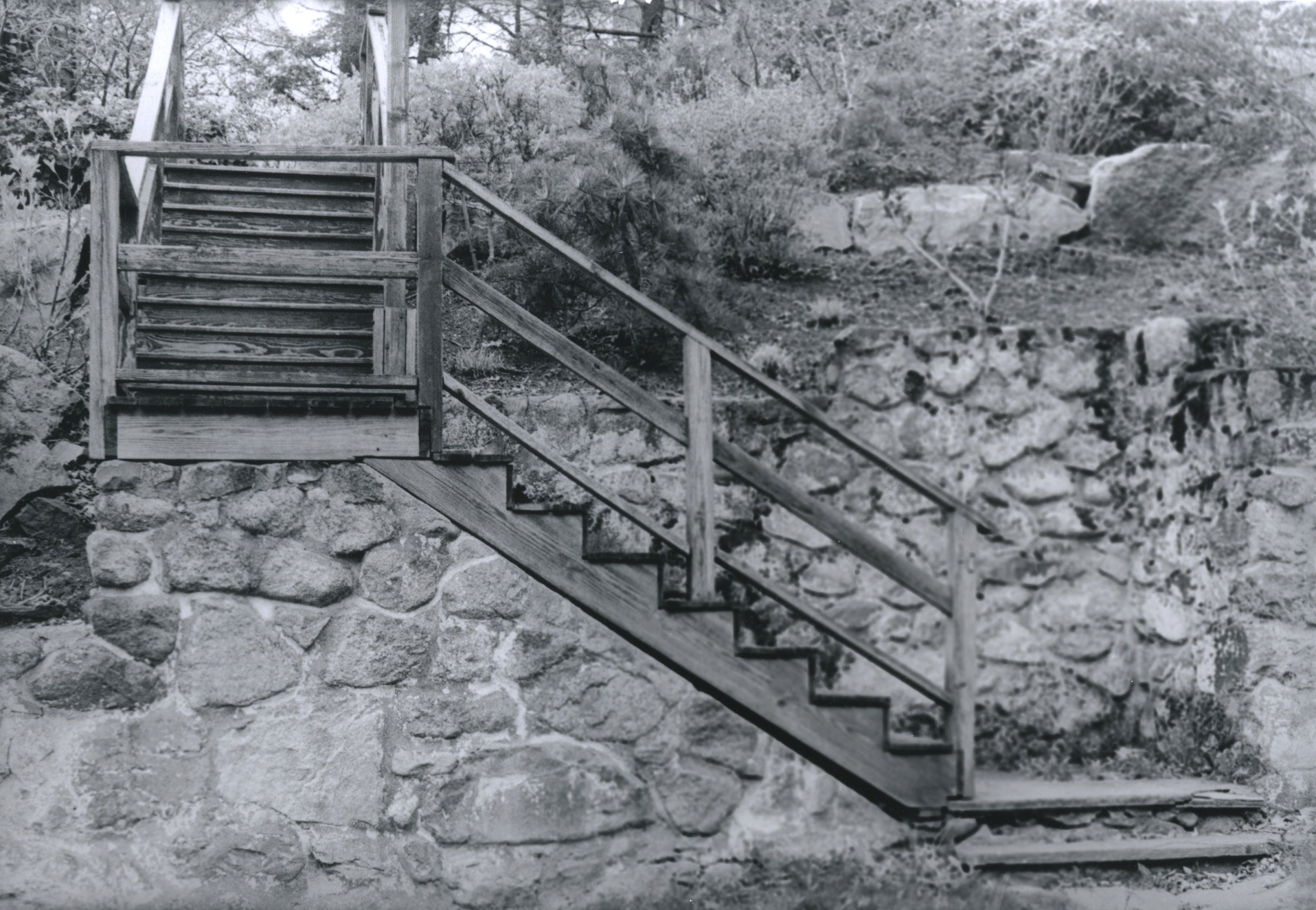 Black and white film print of a set of stairs and a landing leading over a tall retaining wall and onto a hillside embankment
