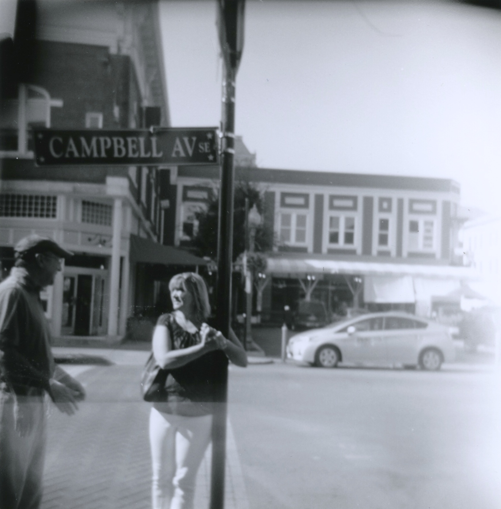 Black and white film print of a man and a woman talking on a street corner while the woman holds onto the "Campbell Av" sign with a dreamy quality from the heavy distortion and vignetting from the plastic camera lens